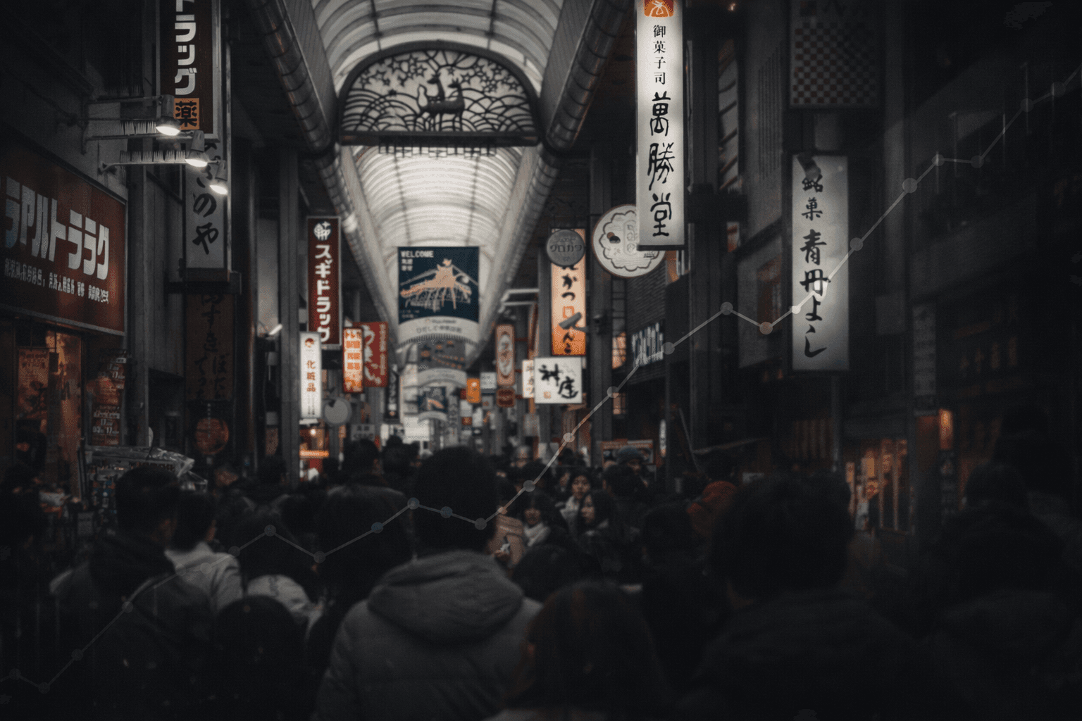 Multidão em corredor comercial coberto no Japão, com lojas e placas iluminadas, representando o cotidiano urbano em meio à pressão do custo de vida.